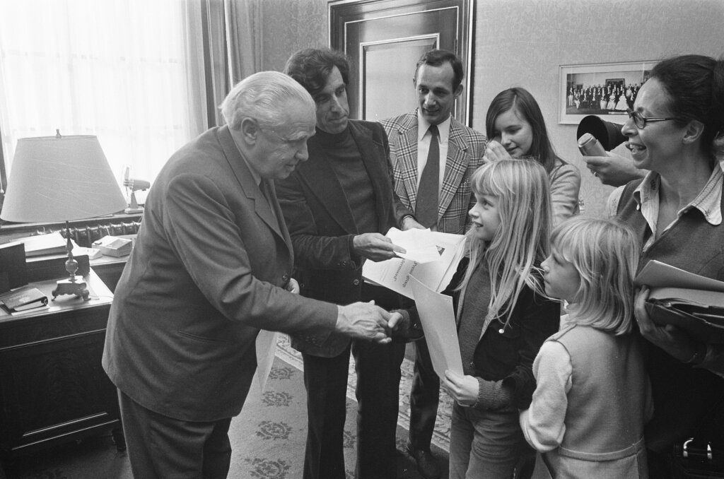 Duas crianças com papeis nas mãos cumprimentam um senhor dentro de um escritório